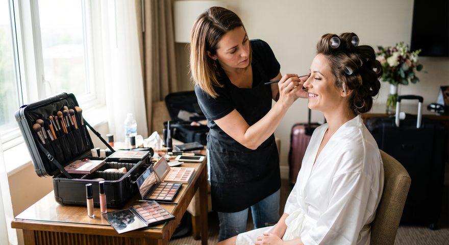A makeup artist in a black apron leaning in with a brush to a seated bride in a white robe with rollers in her hair, in a bright hotel room, an open train case of palettes and brushes on the table beside them