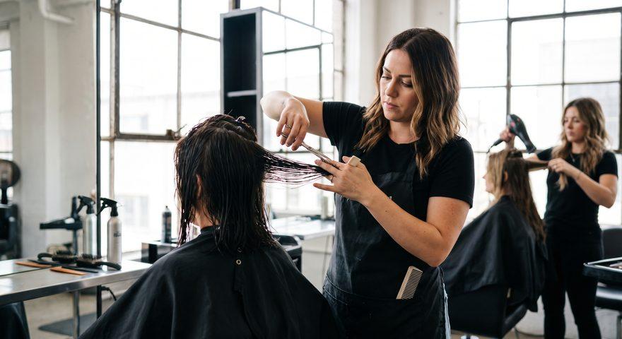 A hair stylist in a black t-shirt and apron cutting a client's wet hair with shears, holding the section between two fingers, in a bright salon where a second stylist is blow-drying a client