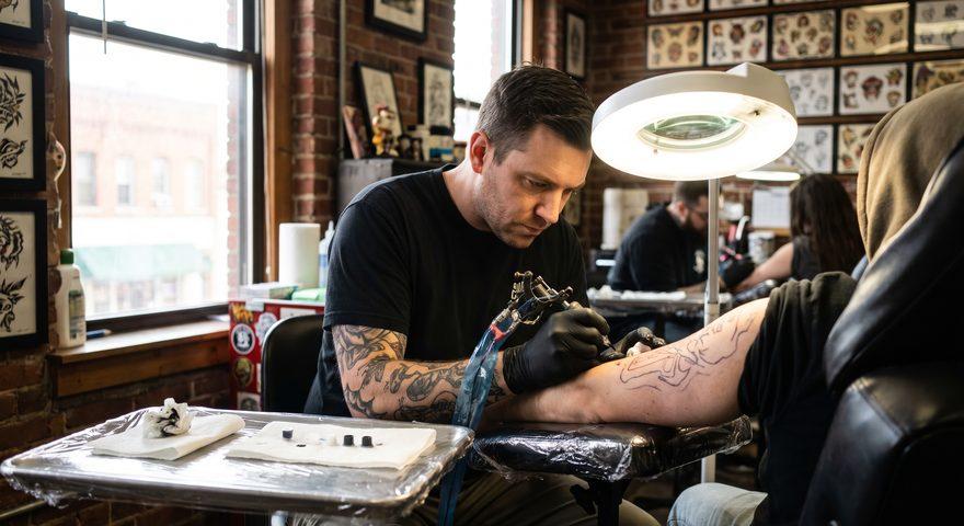 A tattoo artist with tattooed forearms and black gloves inking linework on a client's leg in a brick-walled shop lined with framed flash, under a ring lamp