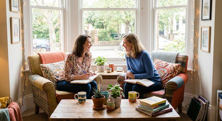 Two women sitting in armchairs by a bay window, notebooks open on their laps, mid conversation